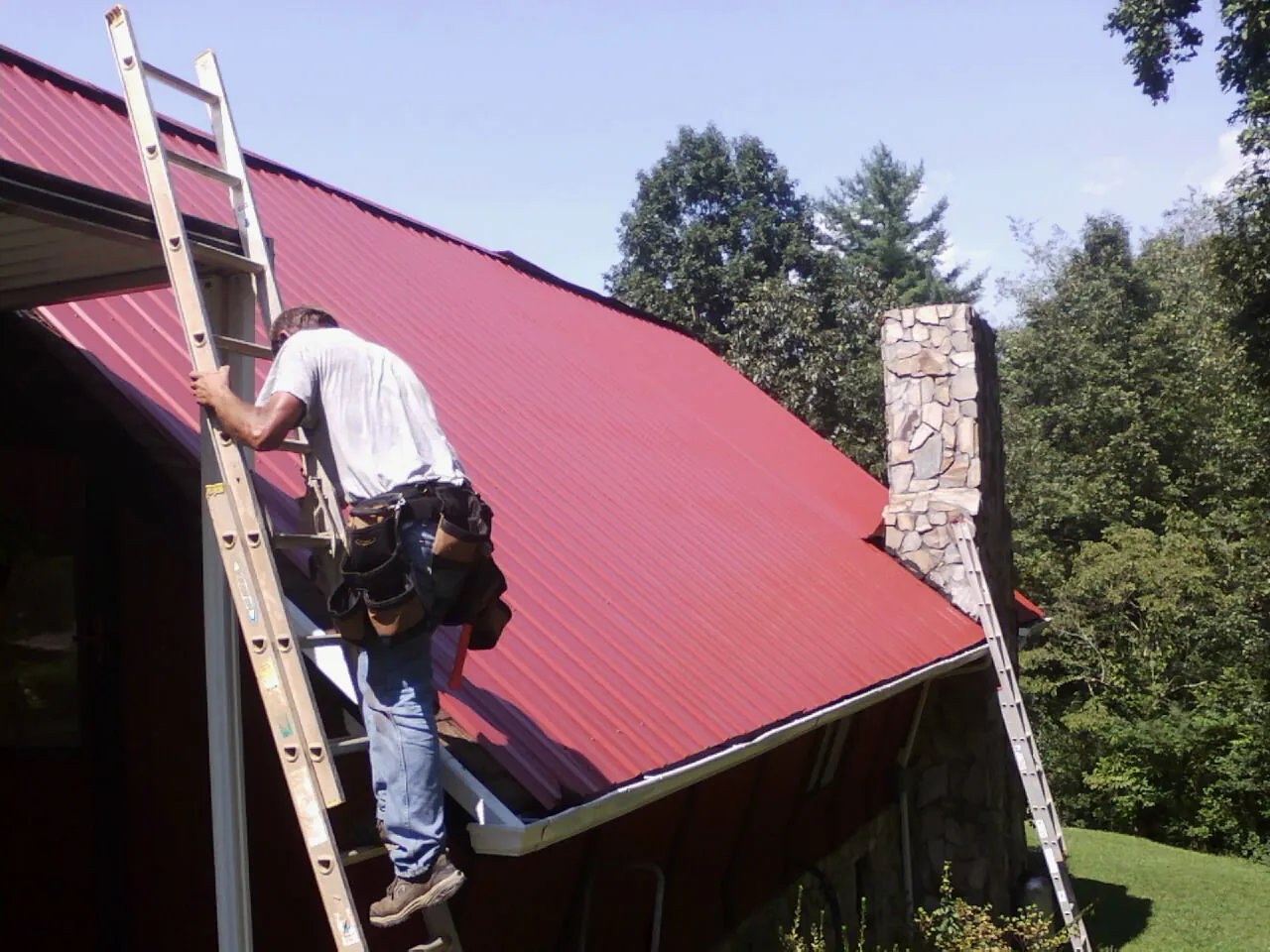 Roofer on a ladder beside a red metal roof and stone chimney — Soffit & Fascia Repair by Roofing Brothers in Cobbtown
